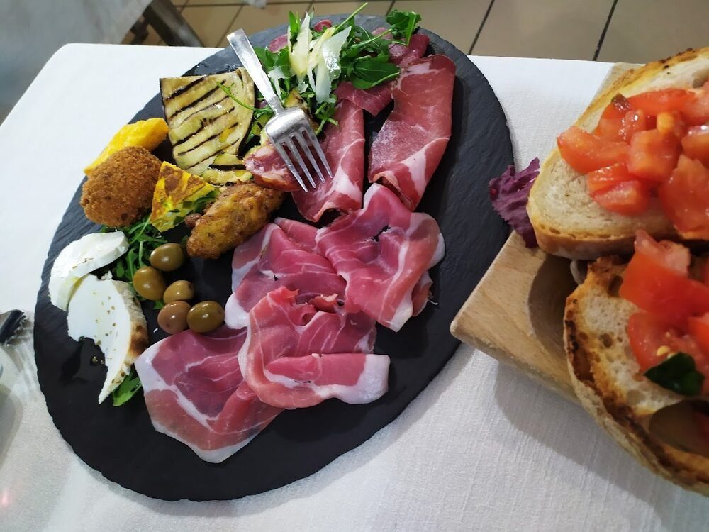 Tagliere di ardesia con prosciutto crudo, coppa, mozzarella, olive, melanzana grigliata e frittura, con due bruschette al pomodoro su un tagliere di legno accanto.