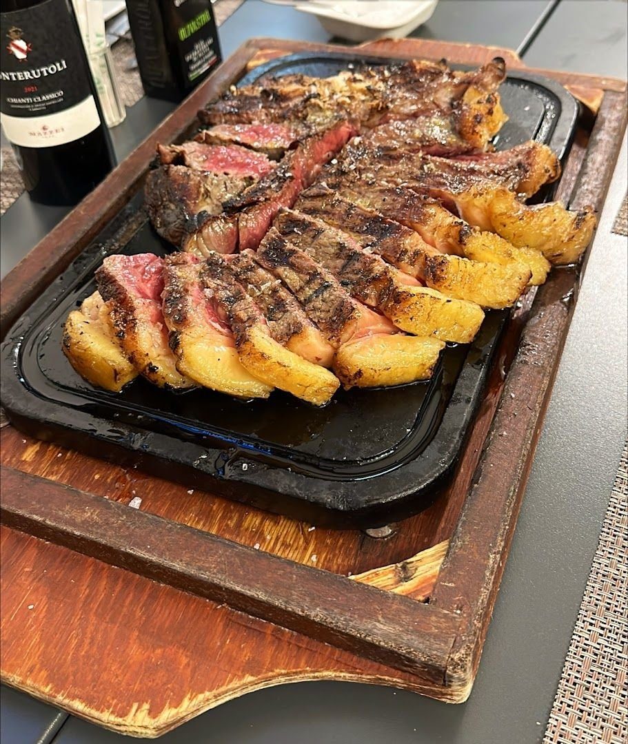 Bistecca alla brace affettata sulla piastra di ghisa dentro il tagliere di legno, con due coltelli da carne e una bottiglia di Chianti Classico sullo sfondo.