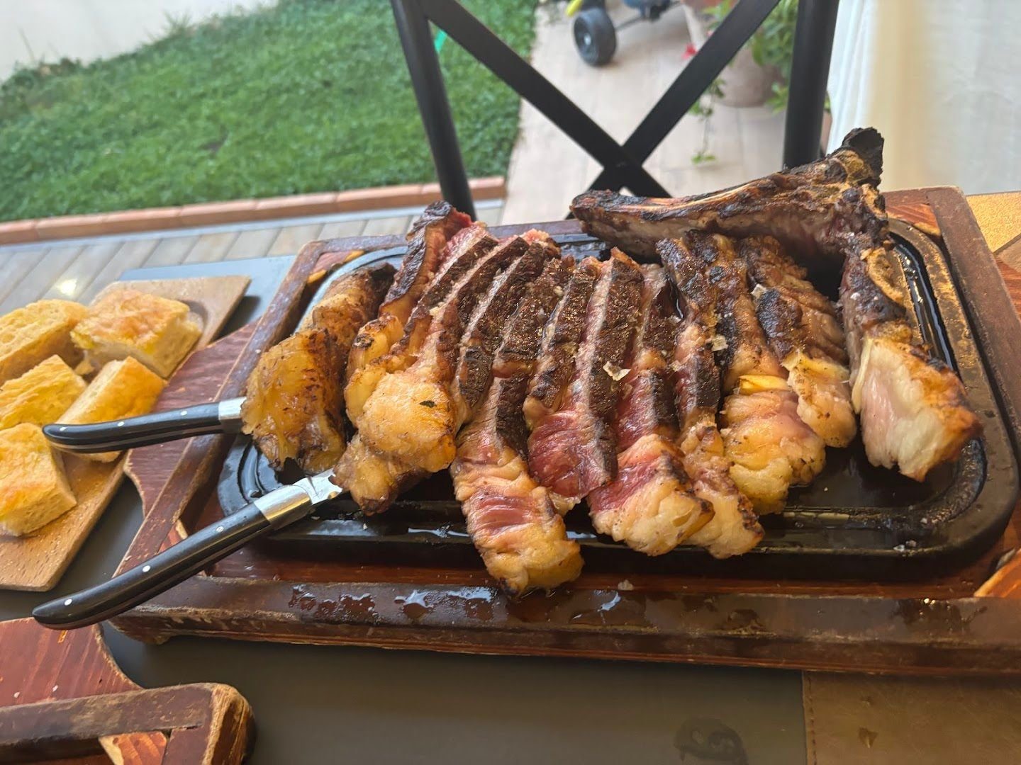 Una bistecca con l&rsquo;osso, cotta alla brace e tagliata a fette sulla piastra rovente appoggiata a un tagliere di legno, con pane abbrustolito accanto.