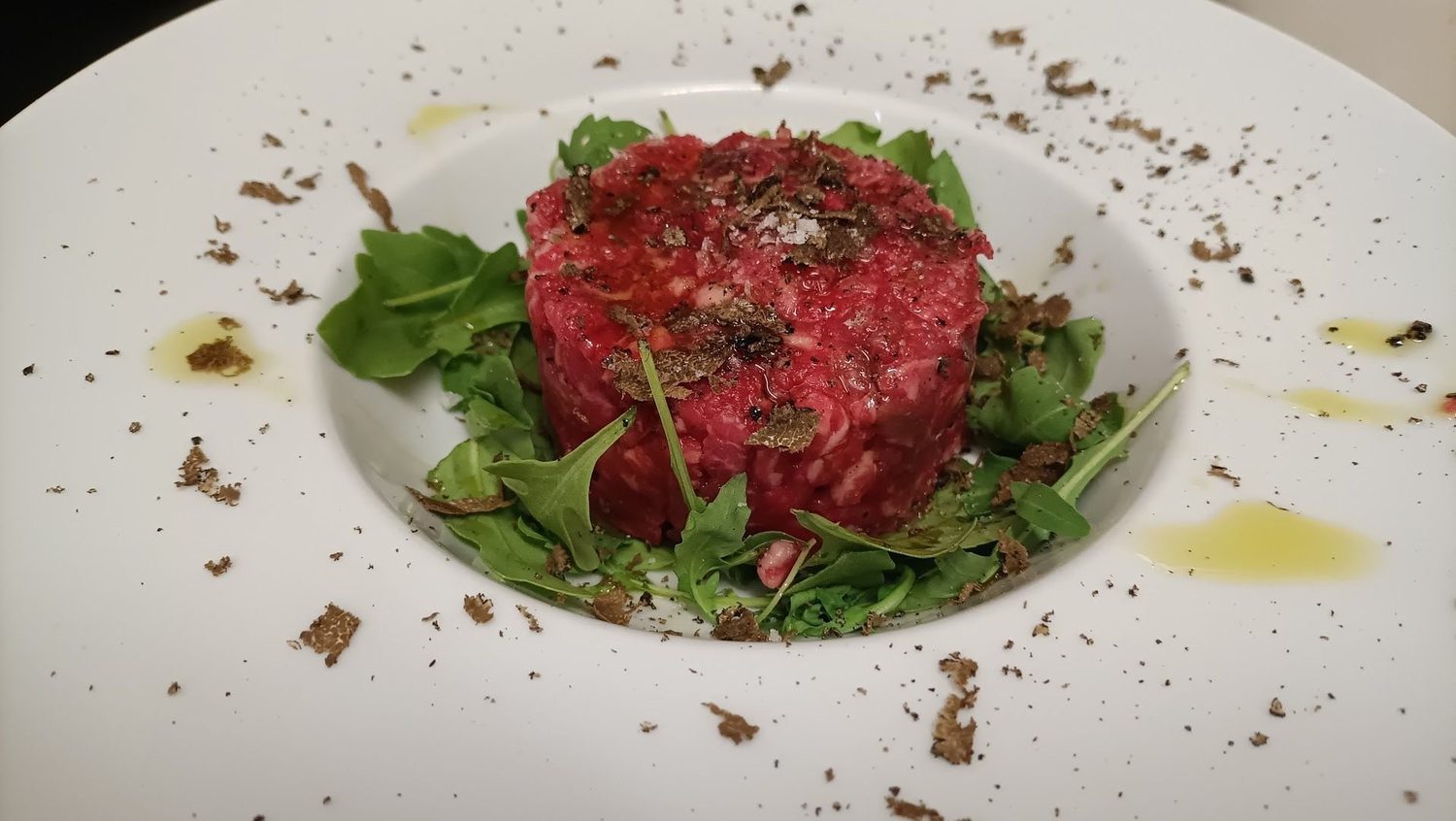 Tartare di manzo su un letto di rucola, con scaglie di tartufo nero, sale grosso e gocce di olio sul piatto bianco.